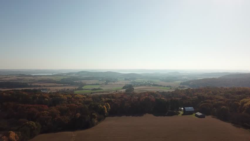 Expansive farmland bordered by vibrant autumn-colored forest, with a barn and hills in the background under a clear sky. Captures a serene rural Midwest landscape.
