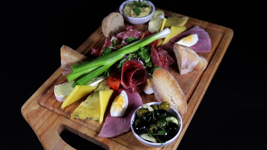 Spinning wooden board loaded with cheese, cold cuts, eggs, bread, and olives.