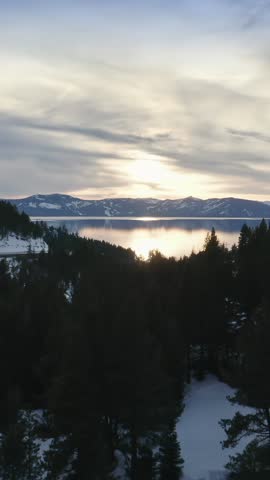 Lake Tahoe, USA, at sunset. The sun reflects off the water, framed by snow-covered mountains and evergreen trees. A road winds along the hillside. 
