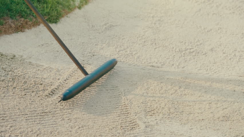 Slow motion closeup of a rake smoothing and grooming sand in a golf bunker. No people visible. Bright sunny day on a well-maintained golf course, capturing the texture and care of course maintenance.