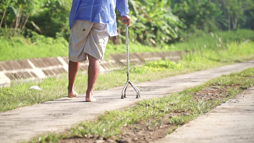 A barefoot elderly man walks slowly along a rural path with the help of a three-pronged walking cane. Captured from behind. Focus on the foot.