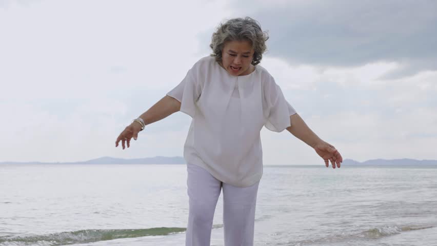 Asian senior woman enjoy kicking the coconut on the seashore and playing with her husband, elderly couple enjoy activity outdoor at the beach, vacation trip after retirement