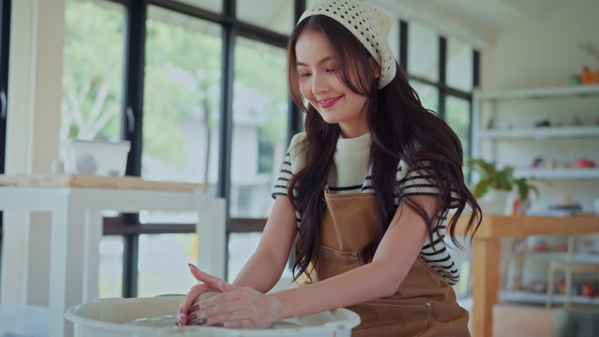 Young asian woman sculpting clay on spinning pottery wheel in ceramic workshop – relaxing creative hobby, art class activity, and handmade design practice in craft studio environment