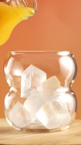Orange juice being poured into a glass with ice on a terracotta background.