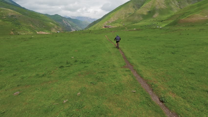 Man riding electric bike along a single narrow trail through open mountain meadow with green hills and scenic peaks in the distance