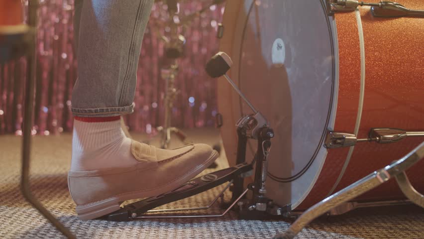 A close-up, low-angle shot of a drummer's foot in a stylish loafer, rhythmically playing the bass drum pedal. The vintage drum and sparkly background suggest a live music performance or band practice.