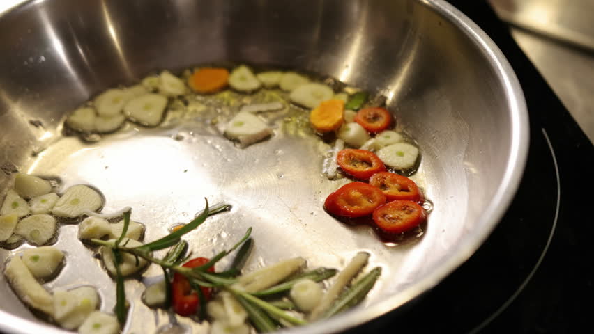 Sautes fresh, garlic slices, frying pan. Close-up sliced garlic, red chili peppers, and rosemary sprigs frying in oil inside stainless steel pan, process of sauteing fresh, aromatic ingredients.