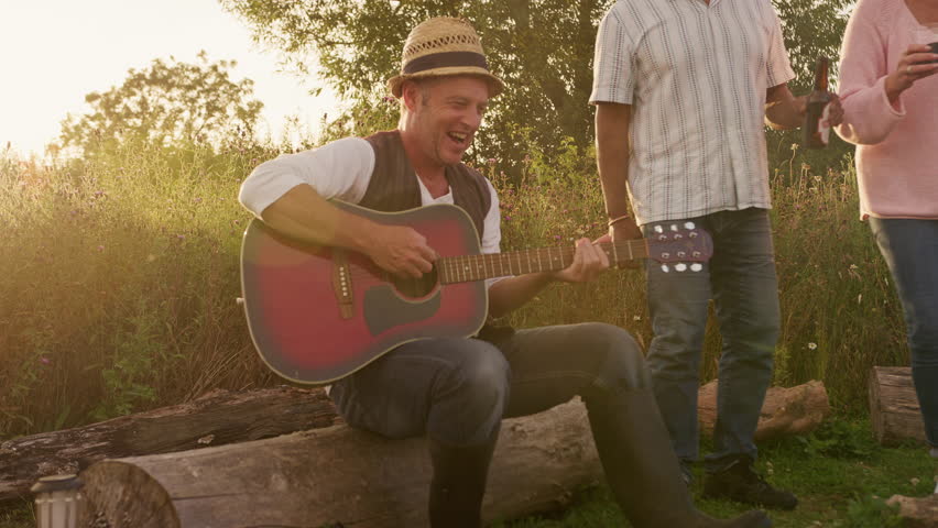 Group Of Mature Friends Sitting Around Fire As They Drink And Sing Songs At Outdoor Campsite