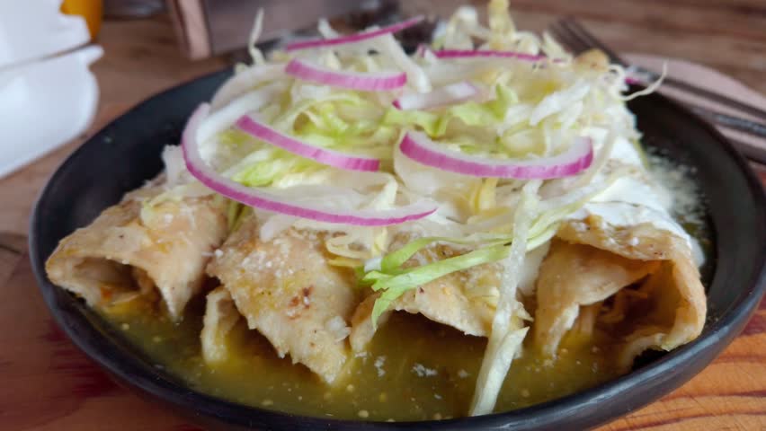 Golden enchiladas with green mole, topped with lettuce and onions on rustic table