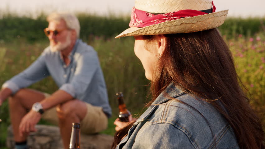 Group Of Mature Friends Sitting Around Fire As They Drink And Sing Songs At Outdoor Campsite