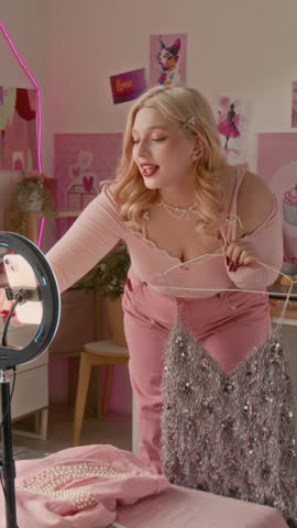 Vertical shot of blonde girl recording beauty fashion blog video with her smartphone, showing her new dress in pink-themed bedroom