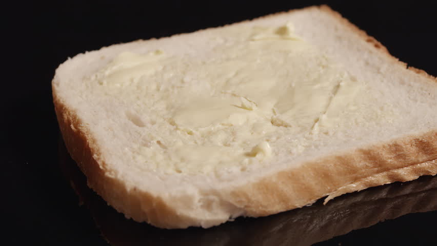 Close-up of a knife spreading vibrant red strawberry marmalade over buttered white bread. Soft top-down shot, black background, highlighting rich textures and glossy jam.