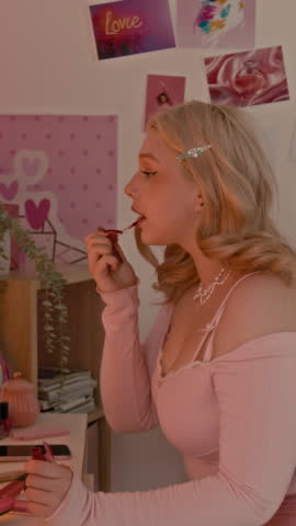 Vertical shot of young blonde girl putting lipstick on while looking in vanity table mirror in her pink-themed room