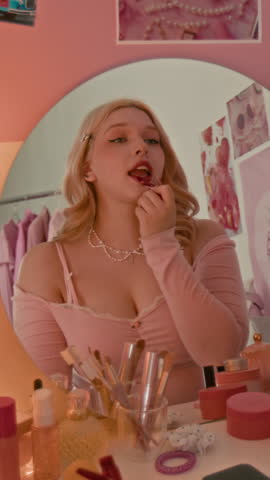 Vertical shot of young blonde girl in pink outfit putting on red lipstick in vanity table mirror reflection