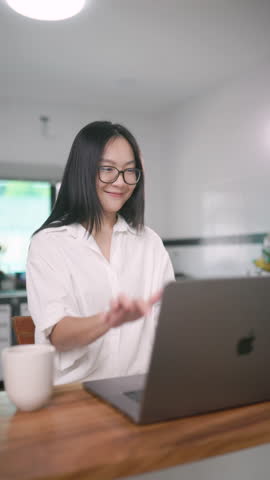 Asian woman smiles during a video call, working remotely from her home kitchen. Real and positive lifestyle of modern remote work.