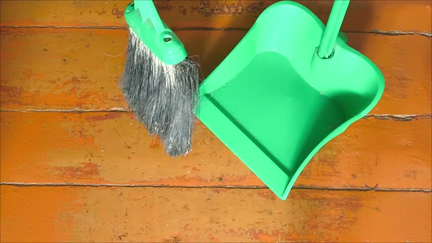 A broom sweeps the trash from the wooden floor into a plastic dustpan. House cleaning. Crumbs, small debris on the floor.