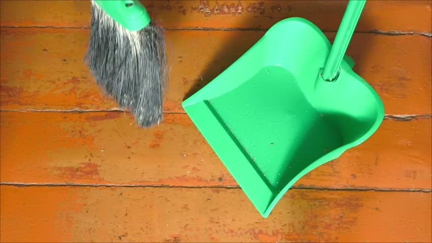 A broom sweeps the trash from the wooden floor into a plastic dustpan. House cleaning. Crumbs, small debris on the floor.