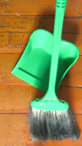 A broom sweeps the trash from the wooden floor into a plastic dustpan. House cleaning. Crumbs, small debris on the floor.