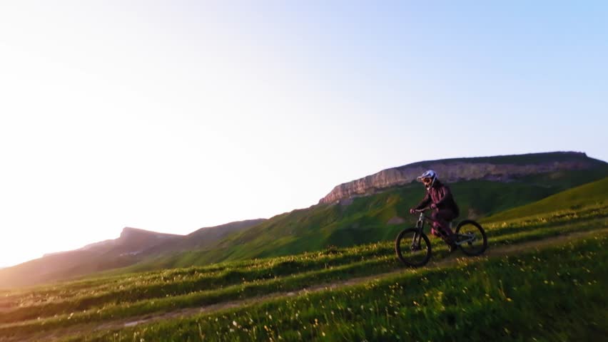 Aerial tracking fpv drone footage of a cyclist doing a wheelie stunt on a mountain bike in a field. Extreme sport activity outdoors in the mountains on a bicycle