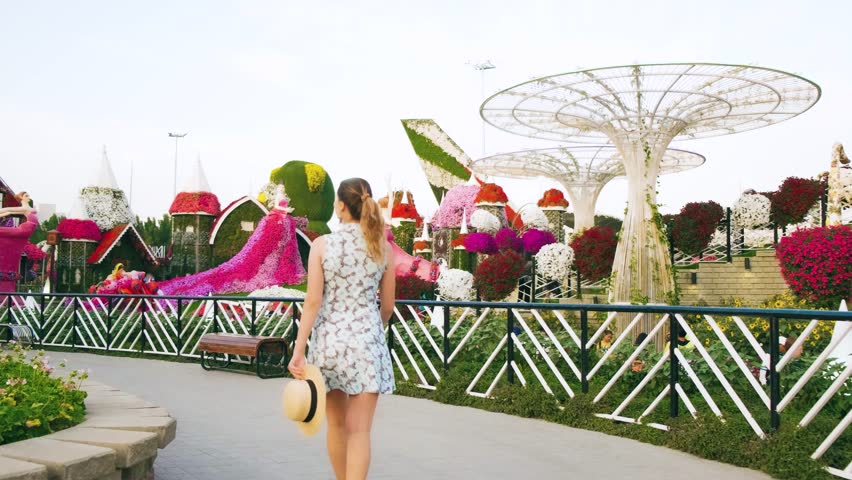 Cheerful happy beautiful young woman tourist walks by alley blooming garden. Young adult girl is spinning dancing,admiring beauty landscape design, colored flowers compositions. Miracle trees. UAE 4k
