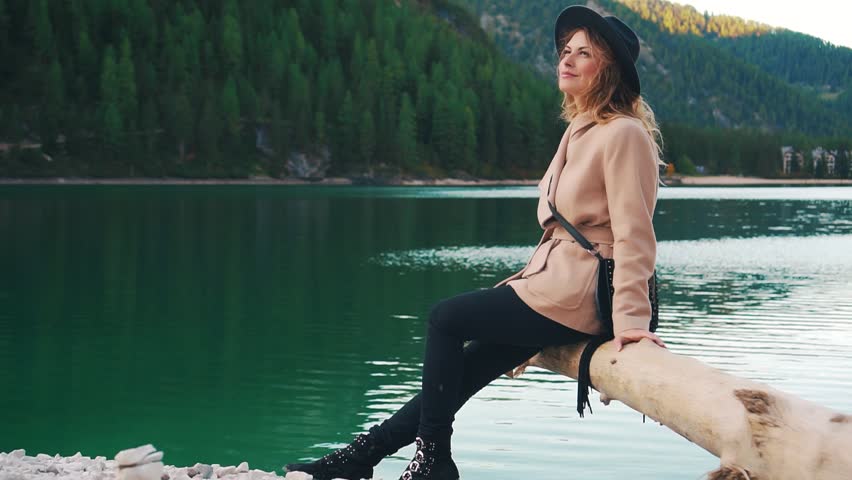 Happy woman tourist in beige nude coat and black hat, inspired girl sitting on log above clear Lake Braies and admiring beauty Italy mountains Dolomites. evening golden hues on turquoise water.