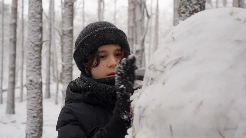 A young boy concentrates on his creative project, carefully shaping and smoothing a snowman. This close-up shot shows his focused effort and diligence during a fun winter activity in a forest.