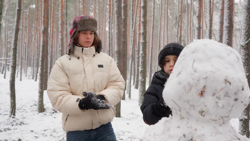 Two brothers work together as a team, adding the final details to their snowman's head. They carefully shape and smooth the snow, finishing their creative winter project in a forest.