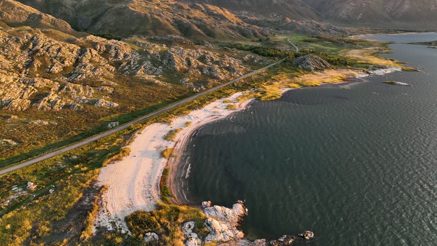 Aerial view of a scenic coastline featuring a tranquil lake, sandy beaches, rugged rocky shores, and majestic mountains under a cloudy sky