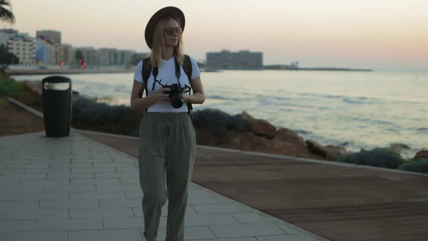 Young photographer walk along wooden boardwalk ocean, capturing golden sunset light with professional camera during scenic seaside travel moment.Travel, blogging, weekend, relax and lifestyle concept