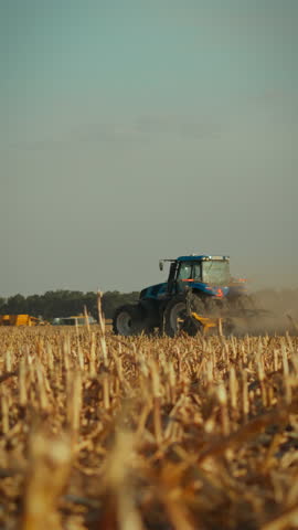 A tractor works diligently in a dusty field during harvest time, gathering crops as the sun sets over rural farmland, showcasing a busy agricultural scene and the beauty of nature