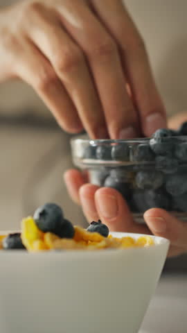 A young woman prepares a healthy breakfast by adding fresh blueberries to a bowl of cereal. This vibrant activity takes place in her kitchen on a sunny day, promoting a healthy lifestyle
