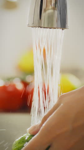 In a cozy home kitchen, a person attentively washes a cucumber under running water, ensuring it is clean and free from dirt before preparing a meal. Fresh vegetables are visible in the background
