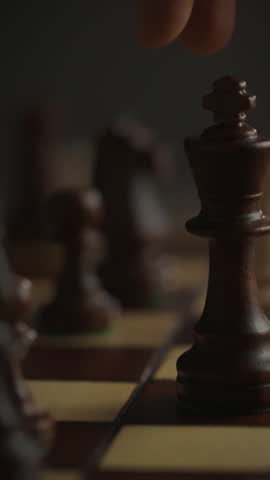 A player focuses on their strategy as they adjust a rook on a wooden chess board during a dimly lit evening match. The ambiance emphasizes concentration and competition between opponents
