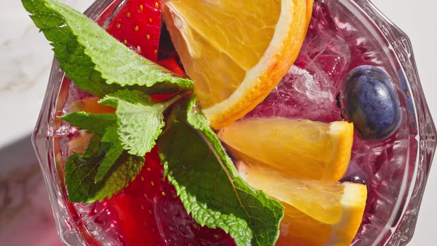 closeup of rotating red sangria glass with fresh fruit pieces, ice and berries, decorated with mint leaf, top view, on sunny light background
