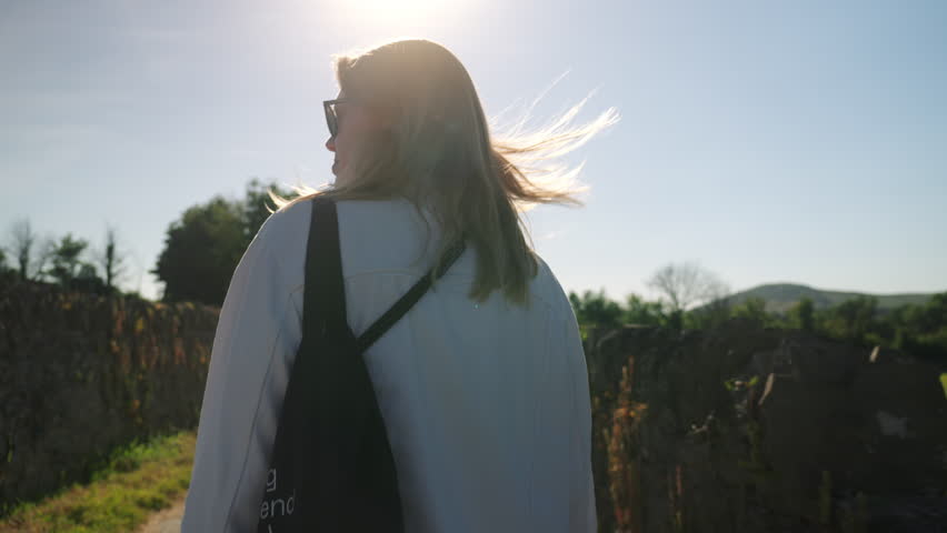 Handheld footage following a pretty blonde in nature with sunlight. Young woman in white jacket walking forward on path in summer.