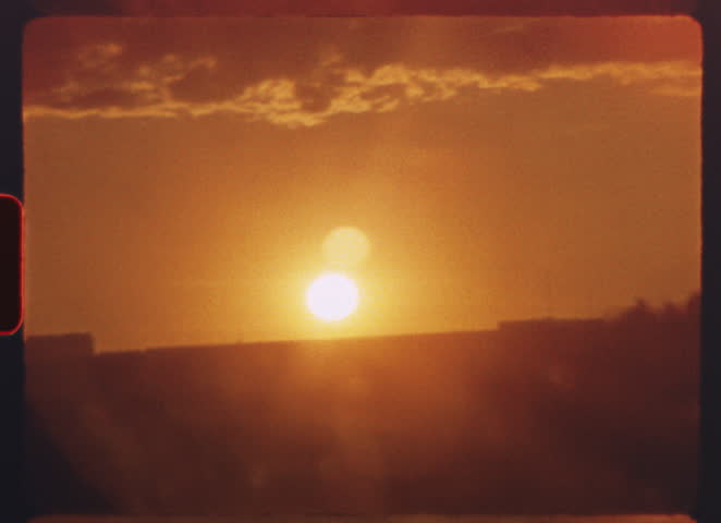 8 mm film - golden sunset illuminating urban skyline, warm orange hues casting soft light through wispy clouds and creating vintage atmospheric cityscape