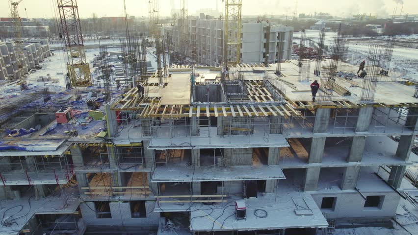 Workers are actively involved in the construction of a multi-story building during winter. Cranes are lifting materials while the site is surrounded by snow and machinery.