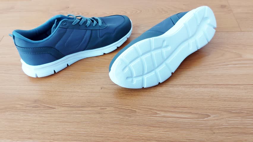 Dark blue sneakers on a wooden floor-one held by hand, the other resting flat. Simple setup highlights everyday style and casual, grounded character