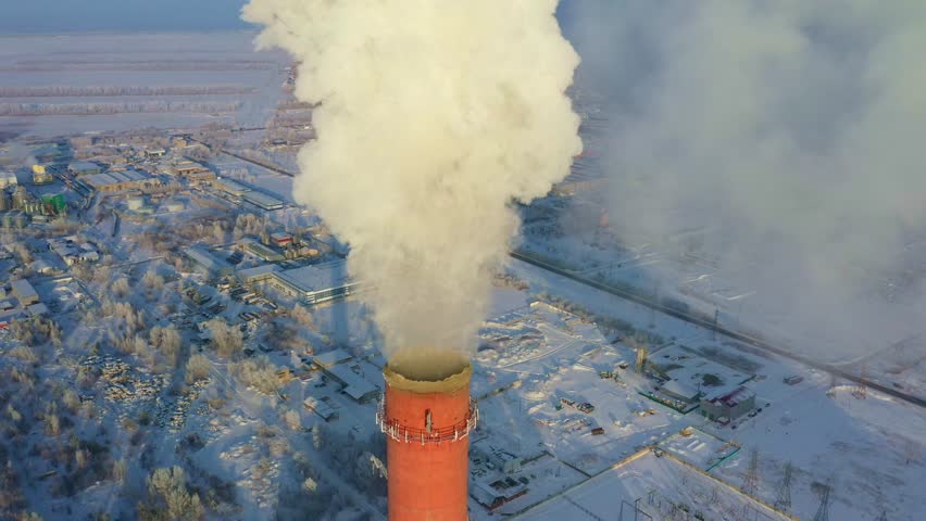 An industrial chimney releases thick smoke into the cold winter air, surrounded by a snow-covered landscape. Nearby buildings and infrastructure are visible in this chilly setting.