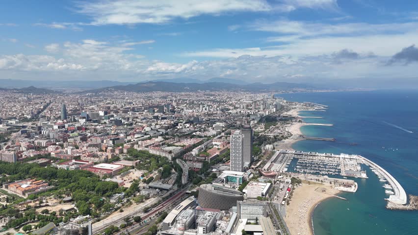 Drone captures sweeping aerial view of coastal Barcelona with Barceloneta Beach, Port Olimpic marina, tall modern building, cityscape, and distant mountain under blue sky on sunny day in Spain