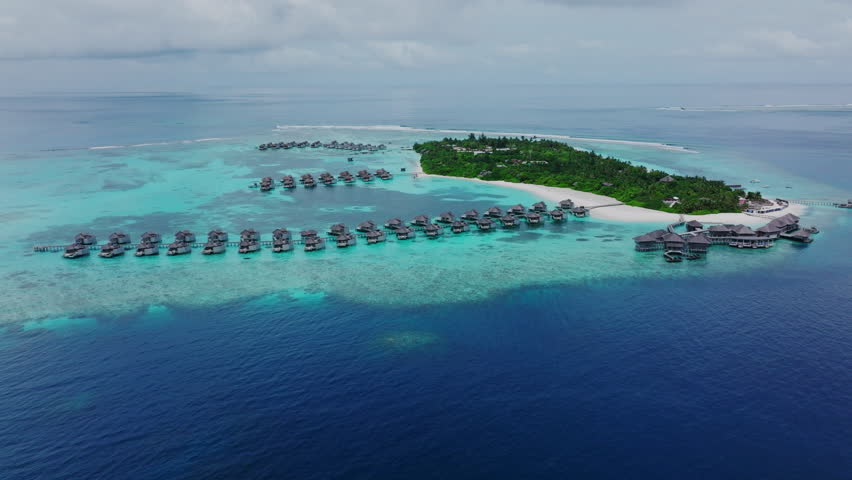 High angle aerial of ring-shaped atoll island in calm Maldives waters with tropical cabana over water bungalows