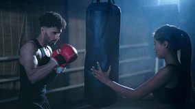 Indian young adult gen z man in sportswear do sport training punching on bag at indoor place. Healthy athletic female boxer teach boxing male wear gloves learn beat exercise workout at dark room - Powered by Shutterstock - Get 15% off with code: PIKWIZARD15