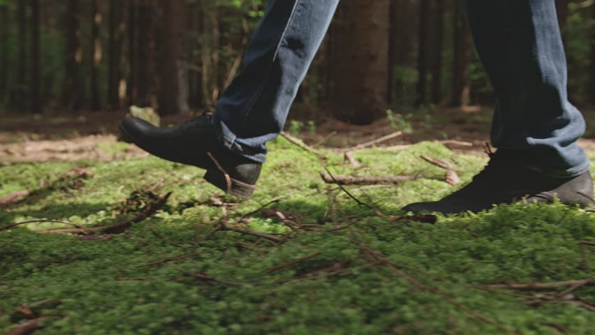 A low-angle close-up shot follows a man's feet in black boots as he walks across a lush, green carpet of moss on the forest floor. Patches of sunlight create a peaceful and serene atmosphere.