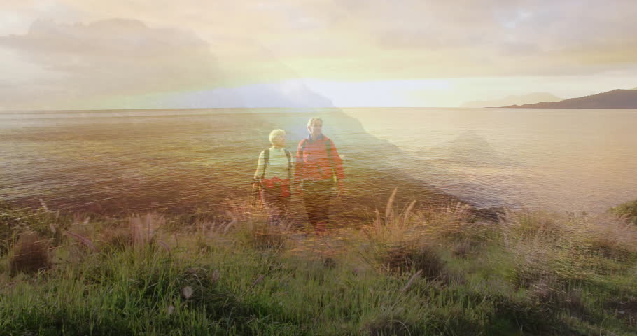 Sun glinting on water spurring senior hiking couple planting pole, walking along dune toward sunset. Adventure, exploration, outdoor, coastline, serenity, silhouette, leisure