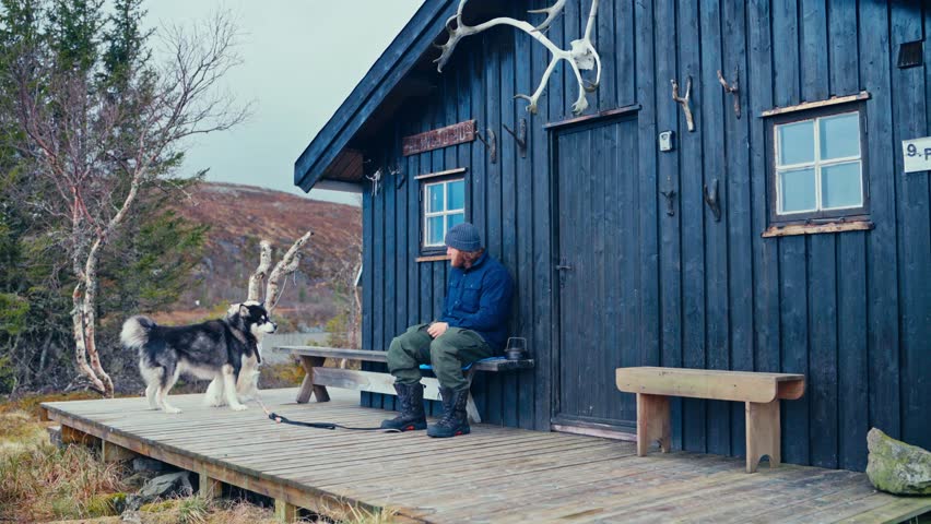 Man In Winter Clothing Sitting On Wooden Porch Of Dark Blue Cabin With Two Dogs. static shot