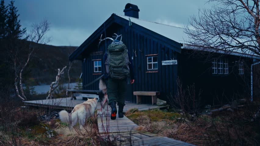 Back View Of Hiker With Backpack And Dogs Walking Towards Wooden Cabin On Cloudy Day. wide static shot