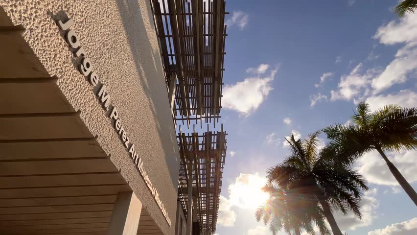 Sunlight streaming through palm trees near the Jorge M. Pérez Art Museum, creates a vibrant scene against a backdrop of blue skies and fluffy clouds in Miami, Florida, United States of America.