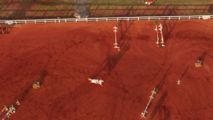 Top down view captures rider and horse jumping an obstacle on red sand in the equestrian arena.