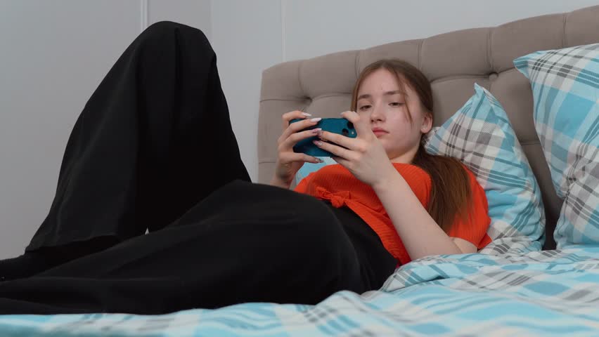 Young teen girl in casual clothes relaxing in bed, browsing smartphone with cheerful expression enjoying quiet leisure at home and playing game on smartphone.