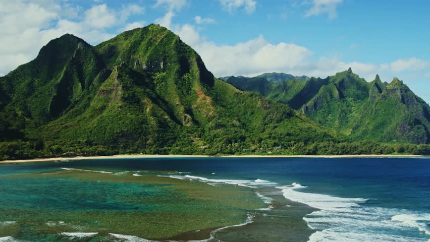 Aerial view flying over coral reef and ocean waves with beautiful green mountains on the North Shore of Kauai, Hawaii